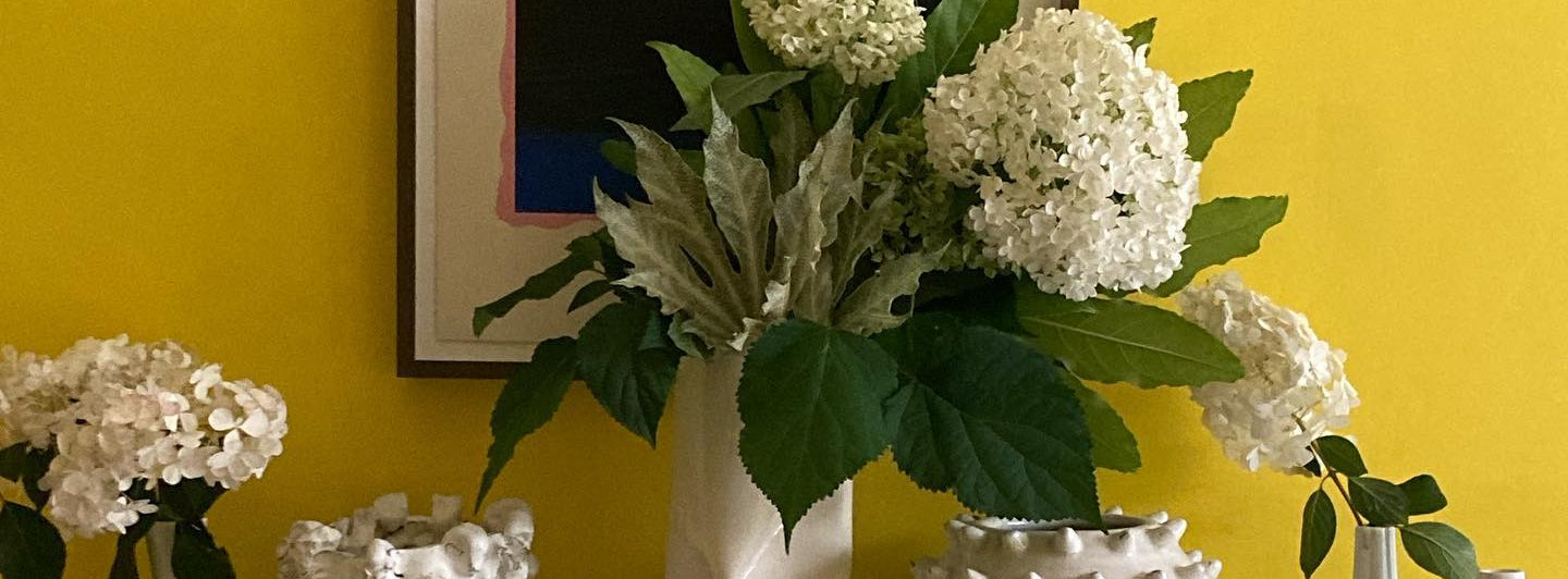 Holland Street. Luxury Linens and Printed Silk Kaftans. Decorative mantelpiece with white vases, flowers, and a colorful abstract painting on a yellow wall.