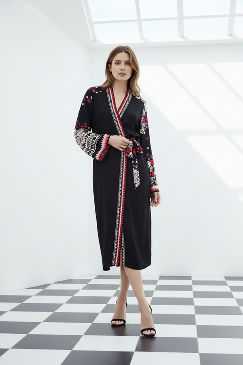A woman models a black long-sleeve kimono-style silk robe with colourful floral print on the sleeves and red, white, and black striped trim along the front opening and hem, standing in a bright white room with a black-and-white checkered floor. From Holland Street London