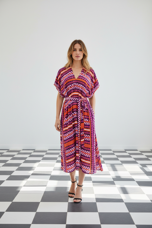 A woman modeling a short-sleeve midi silk wrap dress with a vibrant geometric pattern in purple, orange, red, and white, standing on a black and white checkered floor against a light grey background, from Holland Street London.