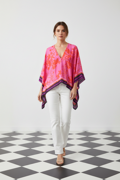 A woman modeling a vibrant pink and orange floral-patterned kaftan top with purple trim, paired with white pants, standing on a black and white checkered floor against a white wall, from Holland Street London