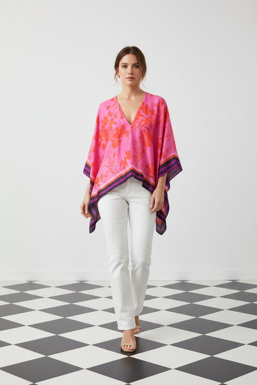 A woman modeling a vibrant pink and orange floral-patterned kaftan top with purple trim, paired with white pants, standing on a black and white checkered floor against a white wall, from Holland Street London