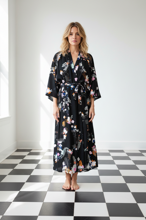 A woman stands barefoot in a bright room with a black floral silk robe featuring pink, orange, and white flowers, set against a white wall and a black-and-white checkered floor, from Holland Street London
